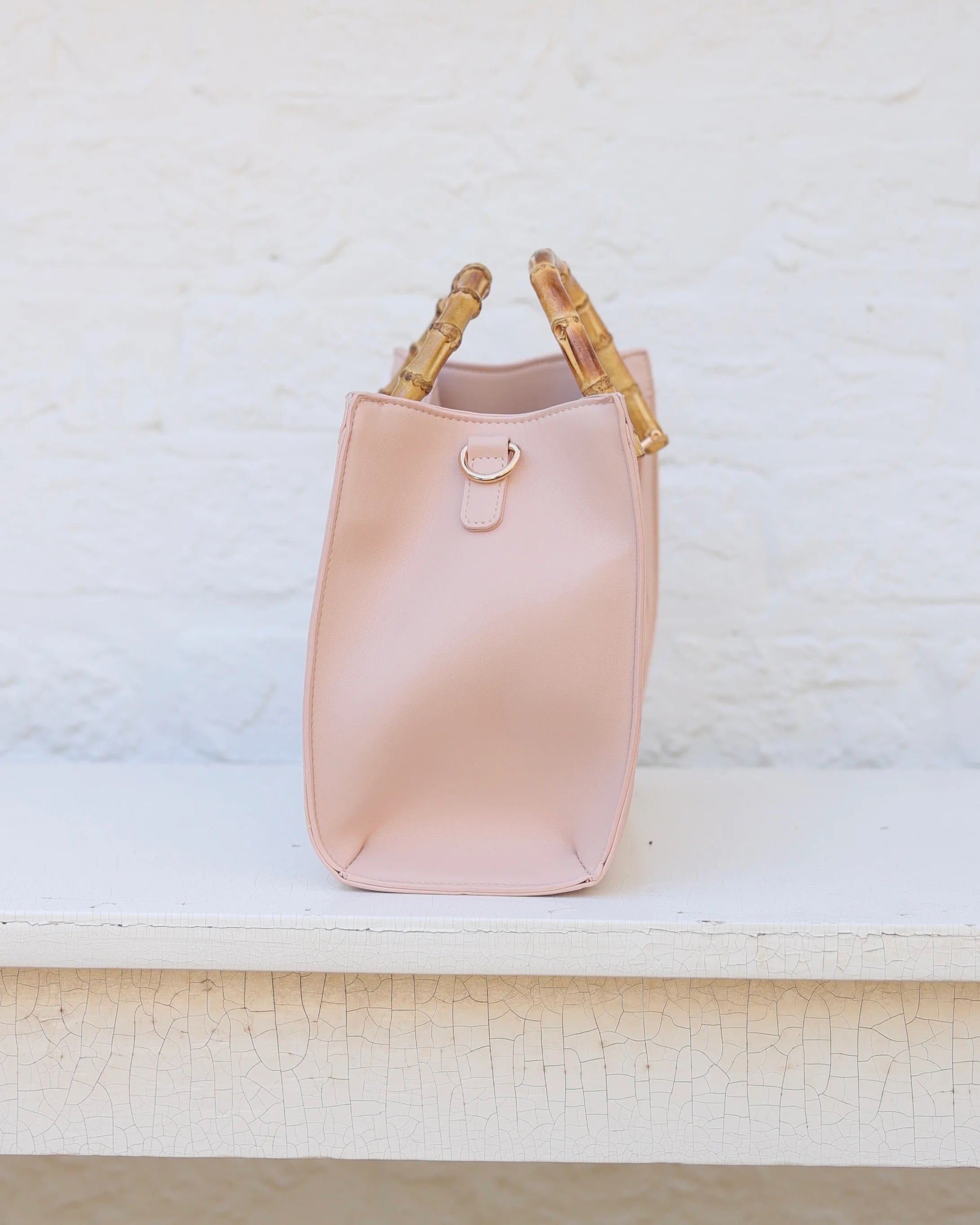 Blush pink handbag with bamboo handles on a white textured background.